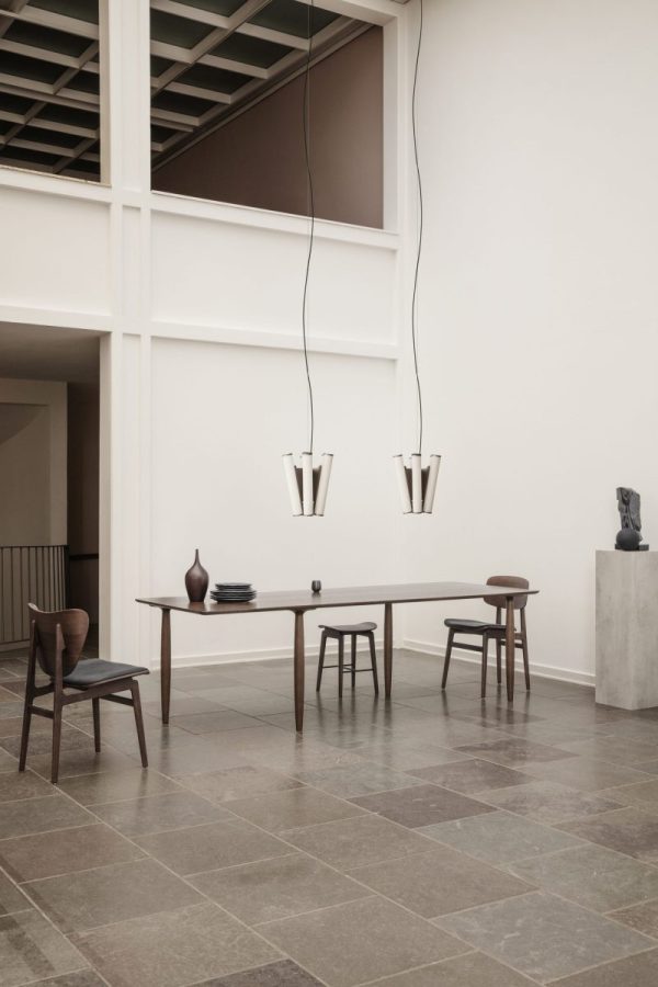 En moderne spisestue med et langt træbord med tre stole og unikke vaser som midtpunkt. Rummet har stengulv, minimalistisk indretning af TomorrowsDesign og store vinduer, der giver rigeligt naturligt lys.