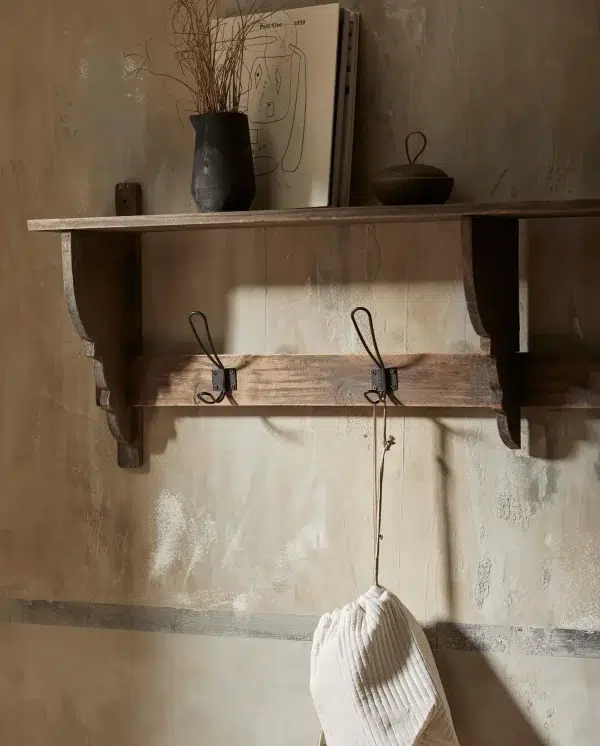 En rustik træhylde monteret på en tekstureret væg med en sort vase, tørrede planter og en bog. Nedenunder holder en jernkrog et hængende hvidt tekstil. Dette interiør komplementerer rummets design perfekt.