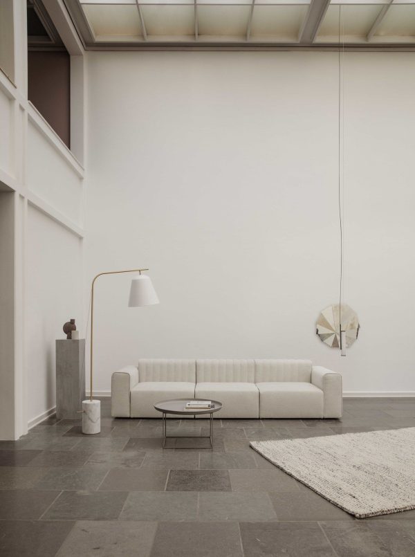 En minimalistisk stue med en hvid sofa, gråt stengulv, et lille rundt sofabord, en NORR11 - Line Two Gulvlampe gulvlampe og et enkelt rundt spejl, der hænger på en neutral-tonet væg.