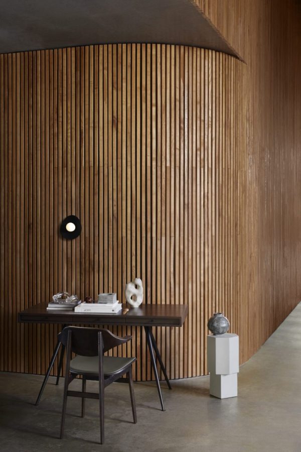 En minimalistisk spiseplads med en trælamelvæg og buet loft, med et mørkt træbord, sorte stole, abstrakte skulpturer og NORR11 - Fuji Væglampe Ø22 lysarmatur. omgivelserne er stilfulde og moderne.