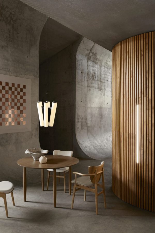 En moderne spiseplads med betonvæg og trelameller. Rummet har et rundt bord med stole, en pendel ovenover og et vægmonteret skakbræt. Minimalistisk design dominerer.