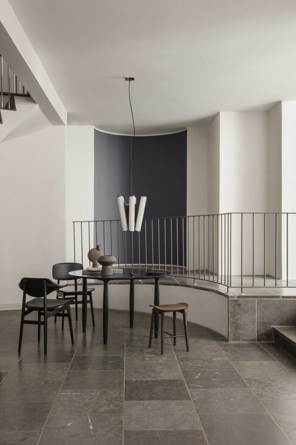 Et minimalistisk interiør i et værelse med en blå accentvæg, med designmøbler inklusive et enkelt sort spisebordssæt, en unik hvid pendel og et stengulv. Området er synligt fra en mezzanin med metalrækværk.