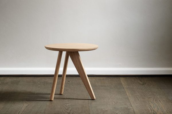 En enkel trætaburet med en rund top og vinklede ben står på et hårdttrægulv mod en almindelig, lysegrå væg, der inkarnerer essensen af TomorrowsDesign.