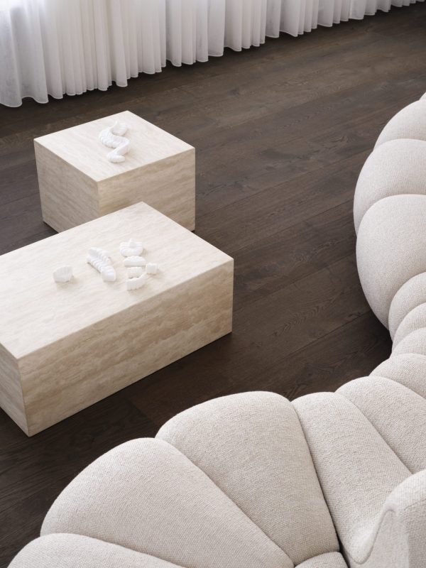 Et moderne spa-rum med to træborde, der viser pænt foldede hvide håndklæder og sæt af spa-sten. En plys, segmenteret beige sofa og gennemsigtige hvide gardiner komplementerer den fredfyldte stemning, forstærket af smagfulde designelementer.