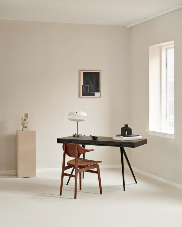 Et værelse i minimalistisk stil med en beige væg med et elegant sort skrivebord med en lampe, bøger og en dekorativ skål, ledsaget af en træstol. Nær vinduet står en piedestal med en skulptur og designmøbler indrammet kunstværk på væggen.