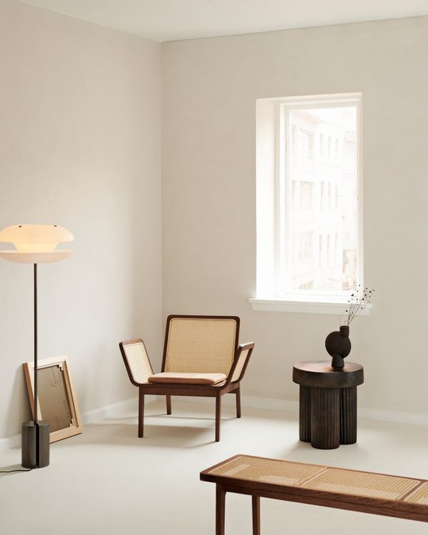 En minimalistisk stue med et stort vindue, der viser udsigt over byen, med en rattanlænestol, en træskammel med en vase på toppen og en gulvlampe ved siden af en lille stak bøger. Dette rum legemliggør TomorrowsDesign med sin innovative tilgang til interiør æstetik.