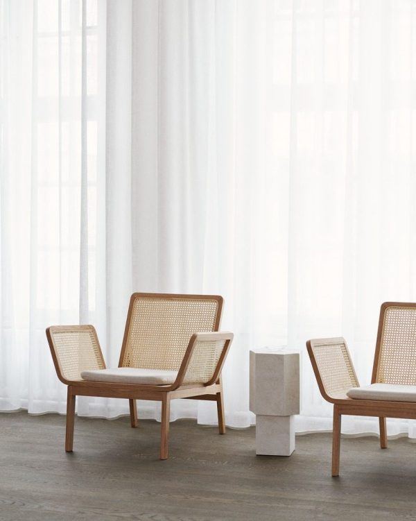 Tre moderne stole med træstel og vævede sæder er arrangeret i et minimalistisk rum med rene hvide gardiner og en hvid marmor piedestal. Rummet, der fremviser udsøgte designmøbler, er badet i naturligt lys.