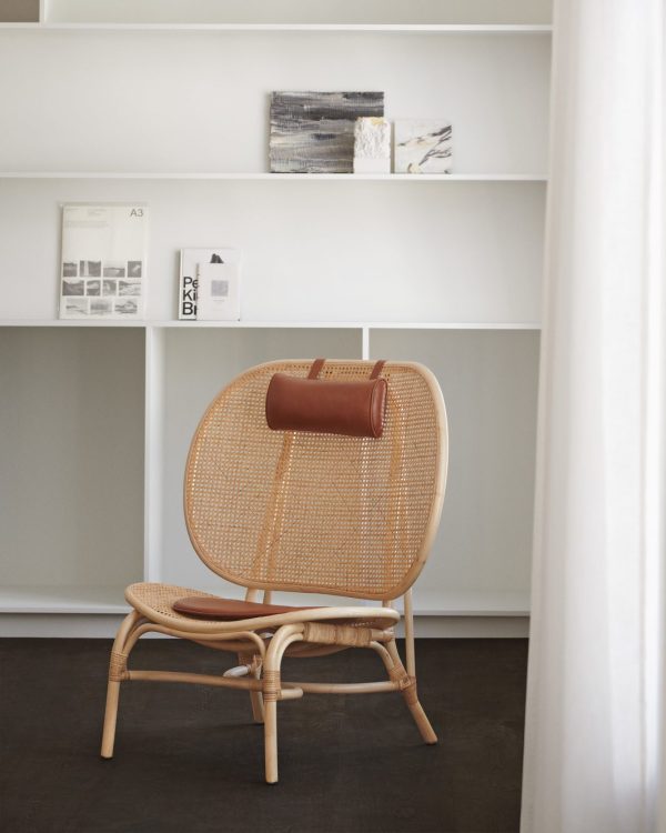 Et minimalistisk interiør med en moderne brun kurvestol med en læderhovedstøtte mod en hvid reol med bøger og dekorationsgenstande, inklusive et lille abstrakt maleri.