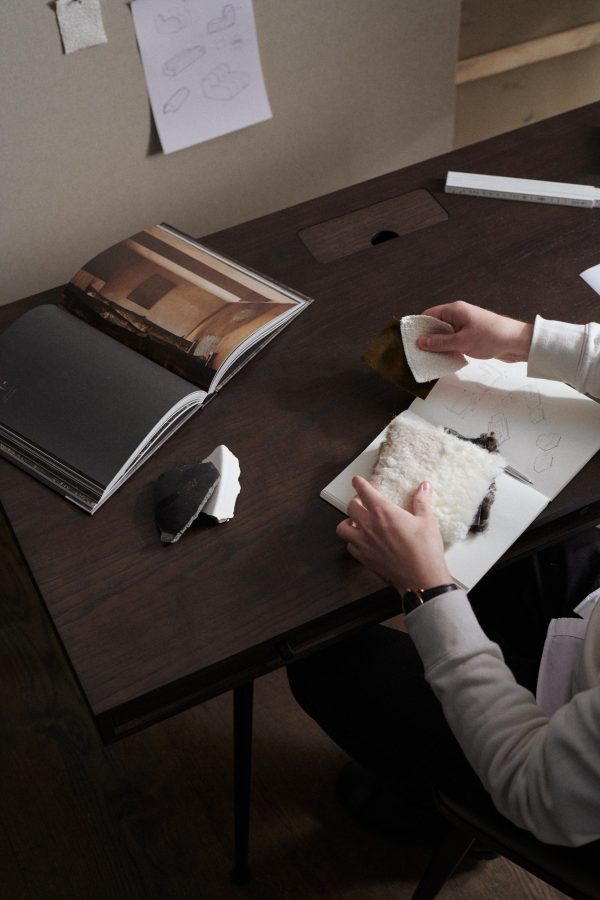 En person sidder ved et træskrivebord og tegner, omgivet af et åbent magasin, tegninger og forskellige teksturerede materialer. Fokus er på den kreative proces med hænder på arbejde i møbeldesign.