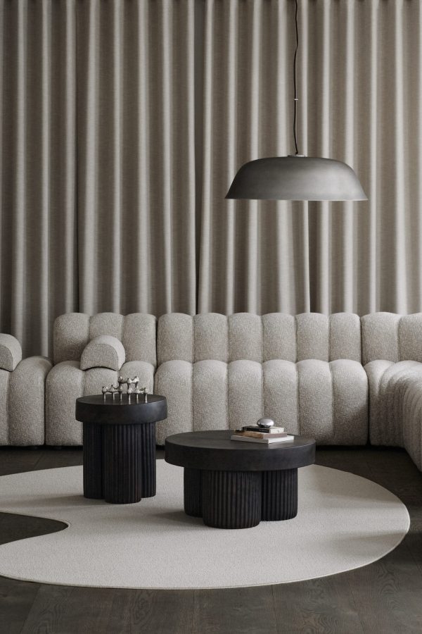 En moderne stilfuld stue med en plys, segmenteret sofa mod et gardinbagtæppe, med to runde, mørke sofaborde på et cirkulært tæppe og en NORR11 - Cloche Three Pendel overhead.
