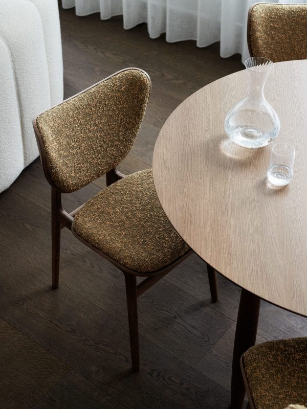 En moderne spiseplads med et rundt træbord med en vase af klart glas og et glas på toppen. To polstrede designmøbler stole med tekstureret stof er placeret nær bordet på et mørkt trægulv.