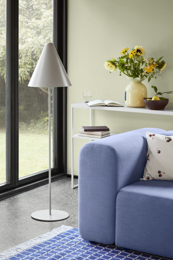 Et hyggeligt hjørne af en moderne stue med en blå sofa med en blomsterpude, designmøbler hvid gulvlampe og en bog på en lille hvid hylde. Der er et lyst vindue og en vase med gule blomster på hylden.