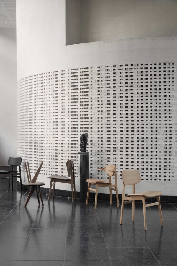 Et minimalistisk spisested med moderne designmøbler inklusiv træstole og -borde på betongulv, der støder op til en stor hvid væg præget af ensartede rektangulære åbninger.