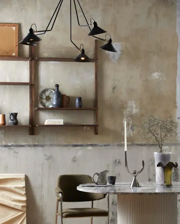 Et rustikt interiør med en nødlidende væg med en træ- og metalhylde prydet med keramik. En mosgrøn stol og marmorbord med pynteting fra TomorrowsDesign under en sort flerarms loftslampe fuldender rammerne.