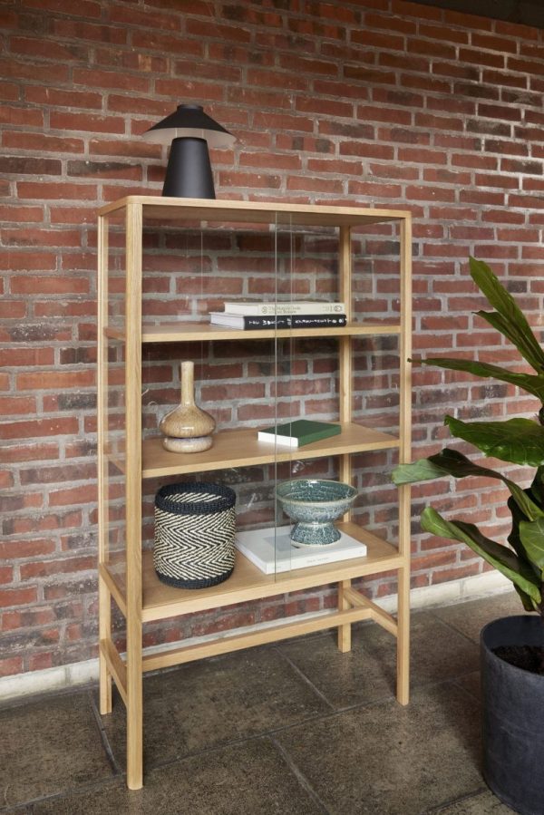 En bogreol i træ med fire hylder mod en murstensvæg. Hylderne rummer bøger, en sort lampe, dekorative skåle og en vævet kurv. En stor potteplante er ved siden af hylden, hvilket forbedrer interiørdesignet.