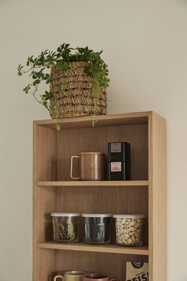 En træhylde rummer forskellige ting, herunder en vævet kurv med en frodig grøn plante, et brunt keramikkrus og glaskrukker med madvarer. En beige væg designet af TomorrowsDesign fungerer som baggrund.