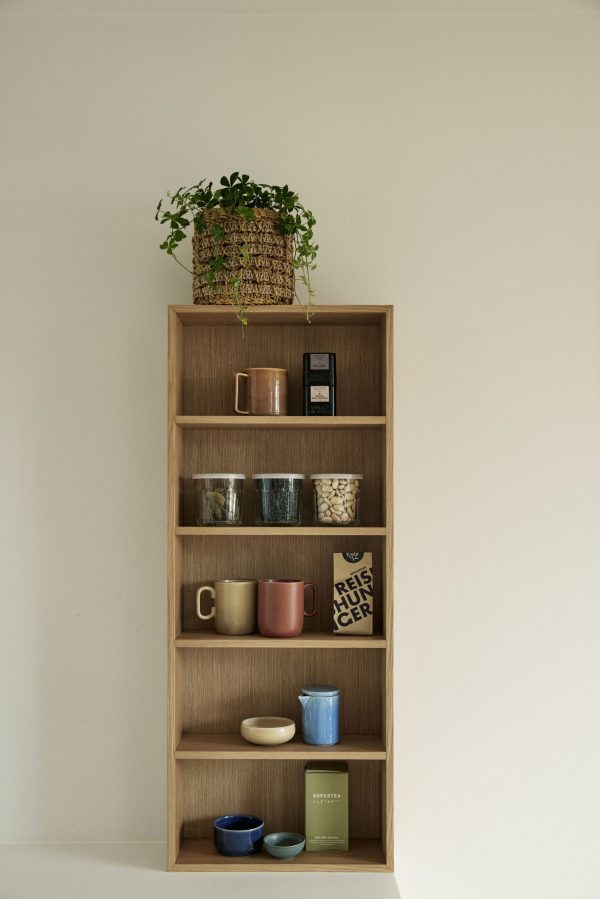 En bogreol i træ mod en beige væg, der viser et design, der rummer forskellige ting, herunder bøger, krus, skåle og en potteplante på øverste hylde.