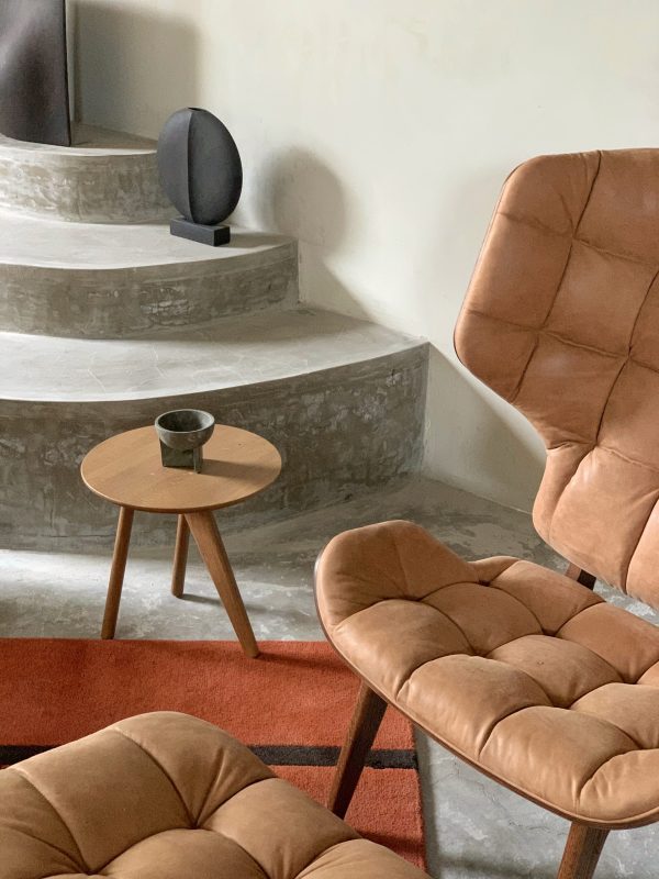 Moderne stue med en brun læderlænestol og matchende skammel centreret omkring et rundt sidebord i træ med en kop på. Et platformtrin og en vægmonteret højttaler er i baggrunden og viser elementer fra TomorrowsDesign.