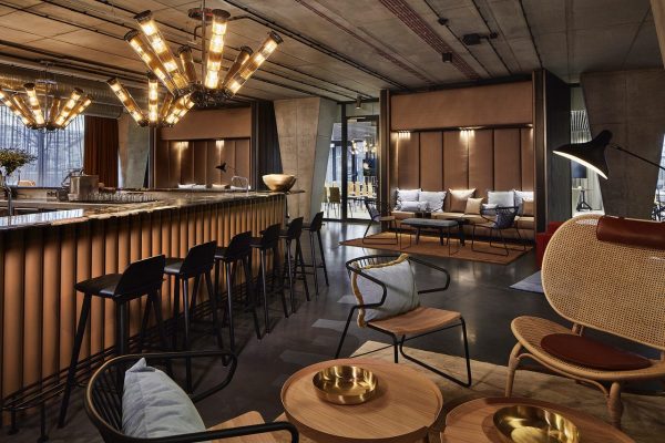 Moderne loungeområde med en bar med høje taburetter, pendullamper og et opholdsområde med plysstole og sofaer, som fremhæves af stilfulde, industrielle interiørelementer.