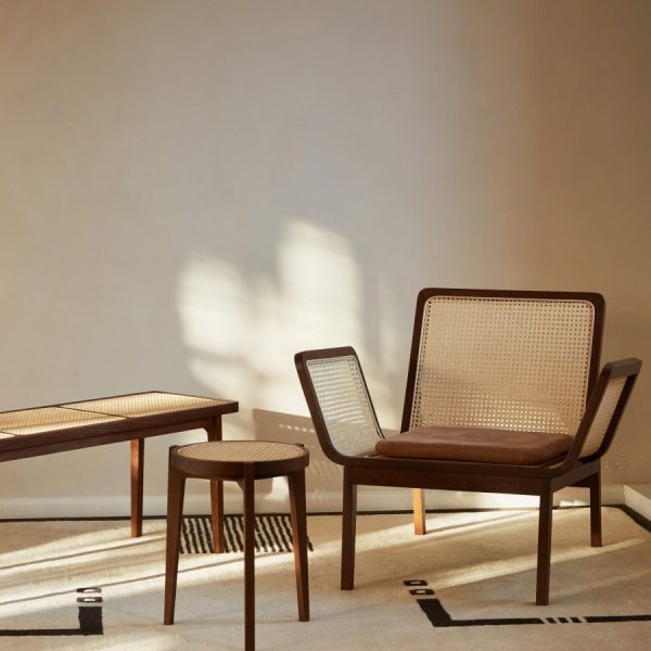 Et minimalistisk værelse med to træstole med stokryglæn og et lille matchende bord, designet af TomorrowsDesign, sat op mod en beige væg med skygger af blade kastet af sollys.