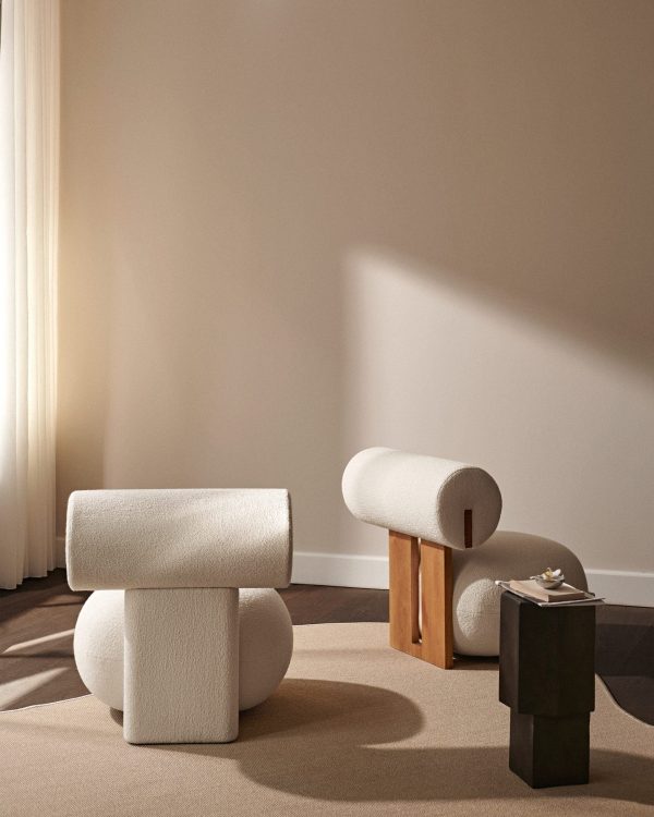 En minimalistisk stue med to unikt formede, plysbeige stole i forskellige størrelser, suppleret med en NORR11 - Quartz Sidebord. blødt sollys filtrerer gennem et rent gardin og kaster blide skygger på et beige tæppe.