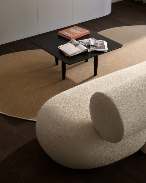 En moderne stue med en unik, cremefarvet cylindrisk sofa, et ovalt sofabord i mørkt træ med magasiner på toppen og et tofarvet møblertæppe på et mørkt trægulv.