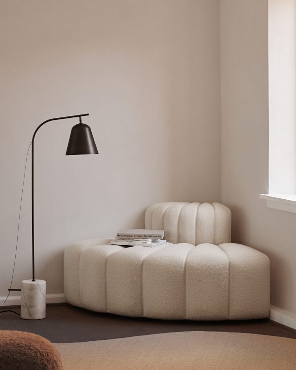 En minimalistisk stue med en cremefarvet, polstret, moderne lænestol med en NORR11 - Line One Gulvlampe lænet henover. et lille sidebord rummer bøger, nær et vindue, der kaster blødt lys.