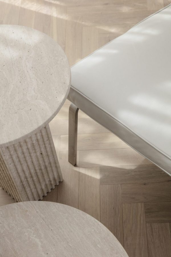 Nærbillede af en moderne stuedetalje med to runde, beige stenborde med ribbede sider, et hjørne af en hvid lædersofa og et sildebenstrægulv fra TomorrowsDesign.