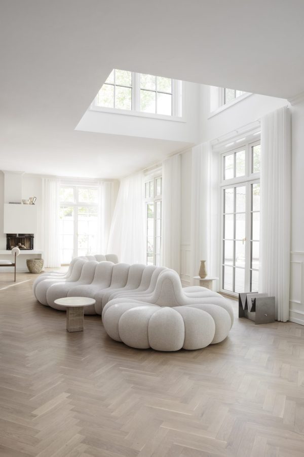 En moderne, rummelig stue med store vinduespartier, hvide gardiner og højt til loftet. Den har en unik designmøbler hvid sofa og et sildebensgulv af træ; naturligt lys fylder rummet.