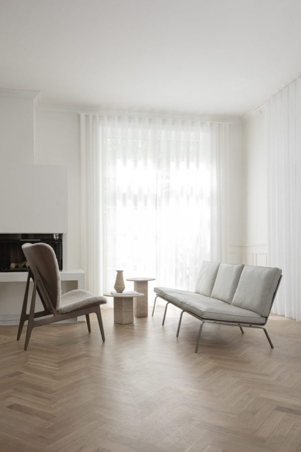 Minimalistisk stue med lædersofa, lænestol, lille træbord og høj lampe. Rummet har parketgulv, hvide vægge og møbler, gennemsigtige gardiner, der slipper naturligt lys ind.