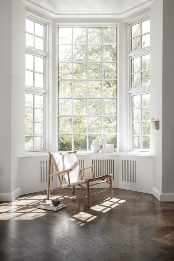 Et lyst og luftigt værelse med store karnapper med udsigt over grønne træer, med trægulv, en minimalistisk gyngestol fra TomorrowsDesign og et lille bord med vase. Sollys bader interiøret og skaber en fredfyldt og indbydende atmosfære.