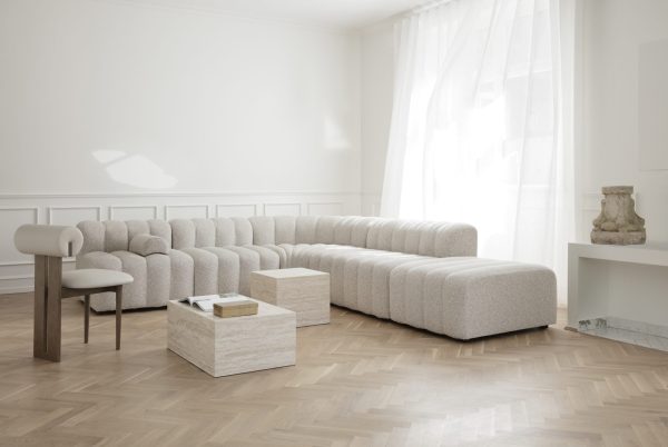 En moderne stue viser en ekspansiv, buet lysegrå modulsofa fra TomorrowsDesign, to små hvide sofaborde, en minimalistisk lænestol og rene gardiner for store vinduer. Gulvene er sildeben af træ.