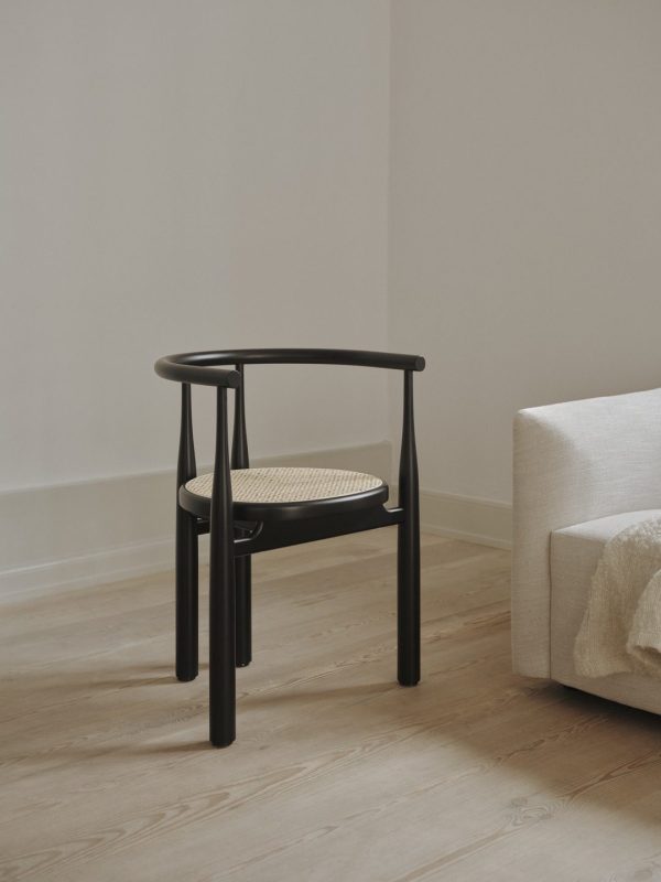 Et minimalistisk interiørdesign med en mørk træstol med et vævet sæde ved siden af en lys beige sofa, placeret på et hårdt trægulv mod en bleg væg.