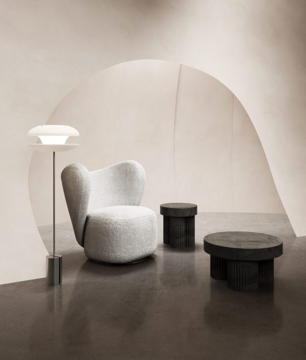 Et minimalistisk interiør med en plys, grå lænestol, to cylindriske borde i mørkt træ og en NORR11 - Yo-Yo Gulvlampe, sat mod en buet beige baggrund.