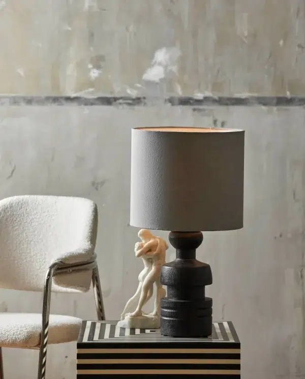 En moderne bordlampe med en unik stablet sort base og en grå skærm, placeret på et stribet sort-hvidt bord. Ved siden af den ses en lille hvid skulptur og en del af en hvid stol mod en struktureret væg. Denne opsætning fremhæver moderne møbler.