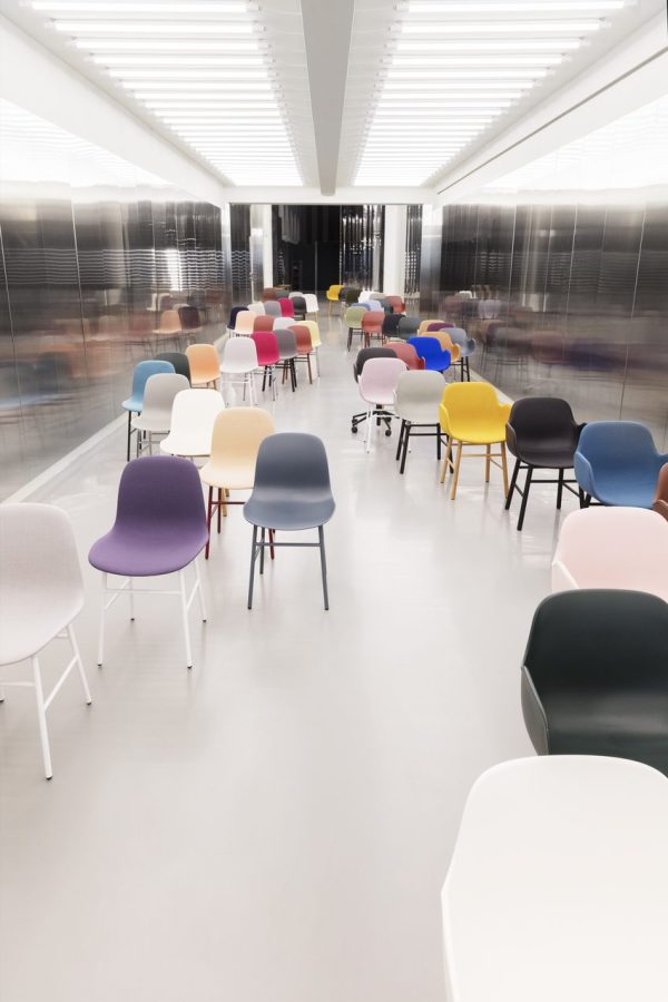 Et moderne værelse med et reflekterende hvidt gulv og metalliske vægge, med en samling af forskellige farvede designstole arrangeret i rækker. Der er en sløret bevægelse af mennesker i baggrunden.