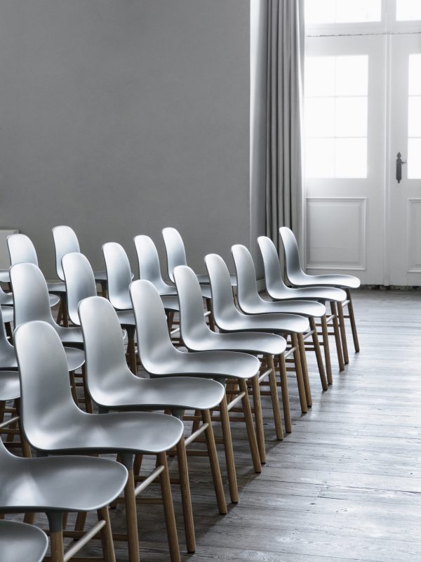 Rækker af tomme hvide moderne stole arrangeret pænt i et rum med grå vægge og et lyst trægulv, nær en lukket hvid dør og et stort vindue dækket af rene gardiner, der viser minimalistisk interiørdesign.