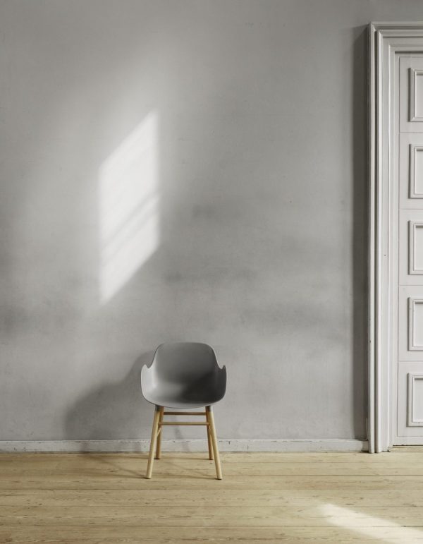 Et minimalistisk værelse med en grå stol mod en hvid væg ved siden af en udsmykket hvid dør. Sollyset strømmer gennem et vindue og kaster et rektangel af lys på væggen og gulvet, hvilket viser TomorrowsDesigns æstetik.