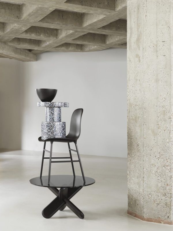 En moderne stol med sort sæde og fod, stablet med glitrende hynder og en dekorativ skål, sat op mod en betonsøjle og loft i et minimalistisk interiør.