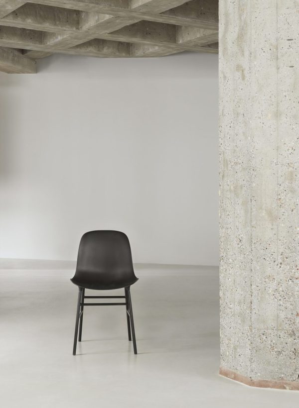 En sort moderne designstol står isoleret på et glat betongulv, støder op til en struktureret betonsøjle og under et bart betonloft i et minimalistisk rum i industriel stil.