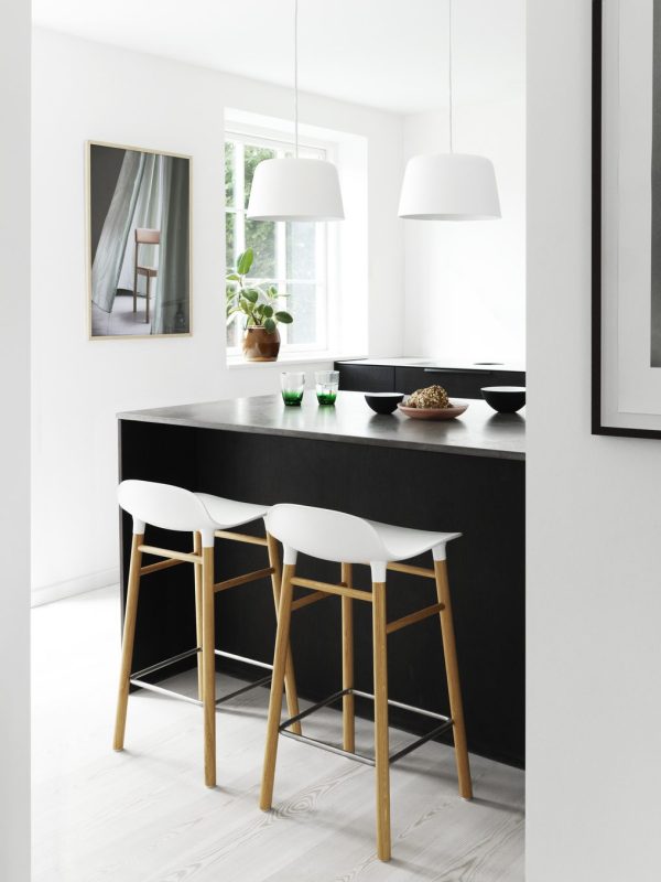 Moderne køkkeninteriør med sort bord og tre hvide barstole, pendellamper ovenover og et indrammet billede på væggen. Der er et vindue med udsigt til grønne områder og små pynteting fra TomorrowsDesign på disken.