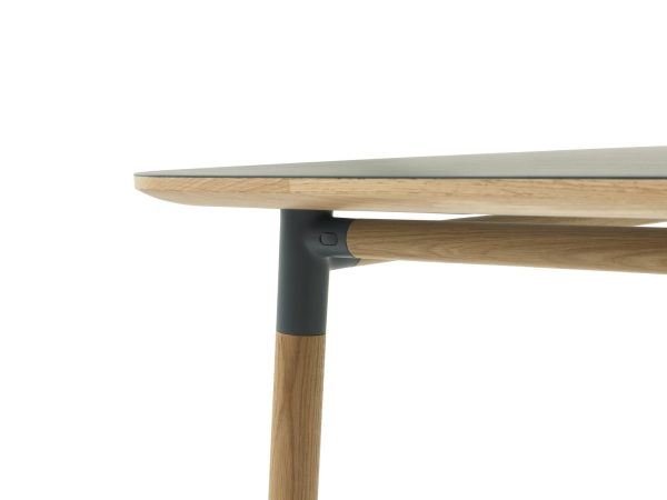Nærbillede af et moderne designmøbler bordhjørne med en lys træplade med afrundede kanter fastgjort til et slankt, mørkegrå ben, mod en almindelig hvid baggrund.