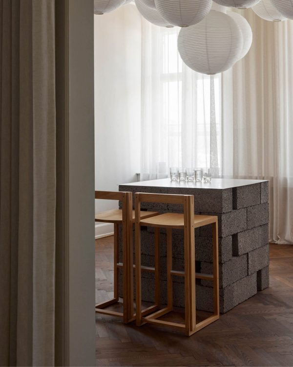 En minimalistisk spiseplads med et bord med en slaggblokbund og en glasplade, ledsaget af KRISTINA DAM - Outline Barstole. rummet er blødt oplyst med store hvide papirslanterner og gennemsigtige gardiner.