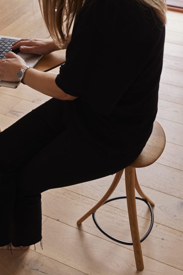 En kvinde i sort påklædning sidder på en træskammel fra TomorrowsDesign, mens hun skriver på en bærbar computer, der er placeret på et lyst trægulv, og hendes blonde hår falder ned ad skuldrene.