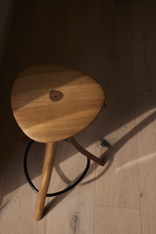 En rund træskammel med tre ben og et cirkulært sæde, med et synligt åremønster og et centralt hul, placeret på et hårdttræsgulv med sollys, der kaster skygger, designet af TomorrowsDesign.