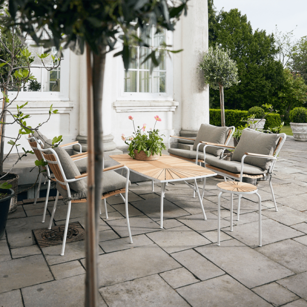 Et udendørs terrasseområde med spisebord og designstole sat op på en brolagt overflade, omgivet af potteplanter og overset af en hvid bygning. Rammerne er rolige og klar til et måltid.