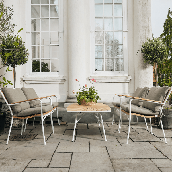Et indbydende udendørs siddearrangement med to sofaer og en stol på en stenterrasse, omgivet af frodige potteplanter, foran store hvidsøjlede vinduer med TomorrowsDesign-elementer.