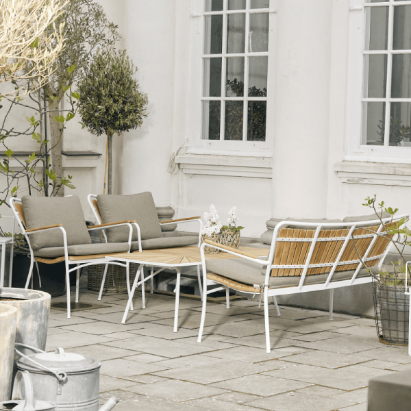 Et hyggeligt udendørs opholdsområde på en terrasse med moderne designmøbler, herunder hvide og træ liggestole og stole, omgivet af potteplanter og træer nær en hvid bygning.