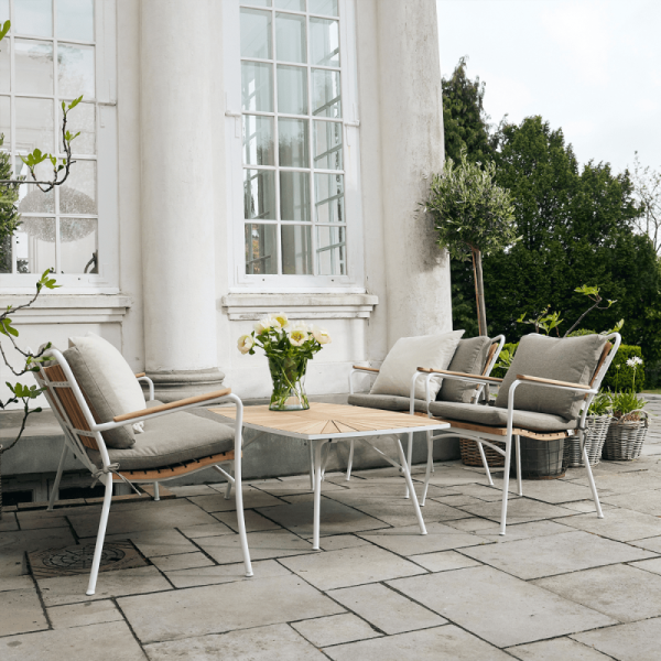 Udendørs terrasse med to polstrede stole og et sofabord, designet af TomorrowsDesign, sat på baggrund af store vinduer og søjler i en klassisk hvid bygning, omgivet af grønne planter.