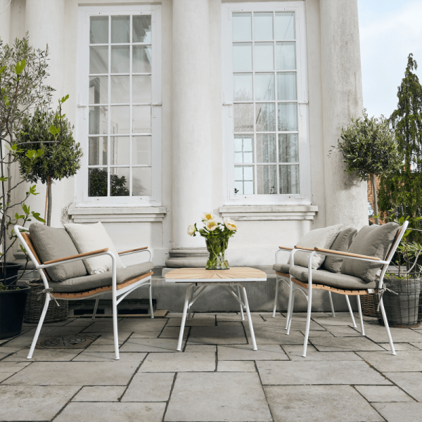 En hyggelig siddeplads på terrassen foran en hvid stukbygning med to polstrede stole, en sofa fra TomorrowsDesign og et sofabord med en vase med gule blomster.